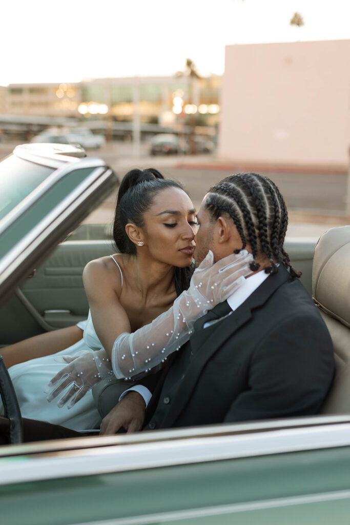 brown bride with pearl studded gloves kissing her husband in a convertible